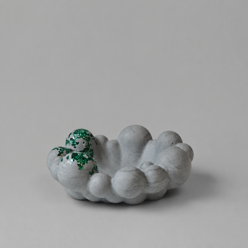 A small sculptural bowl of matte gray stone-like material, with a textured green accent on one side, photographed on a plain light gray studio background.