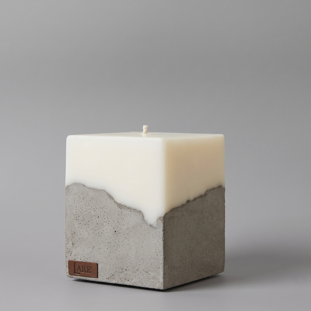 A square pillar candle with a dual-texture design, featuring a smooth soy wax top half and a textured gray concrete bottom half, with a small brown leather brand tag LARE