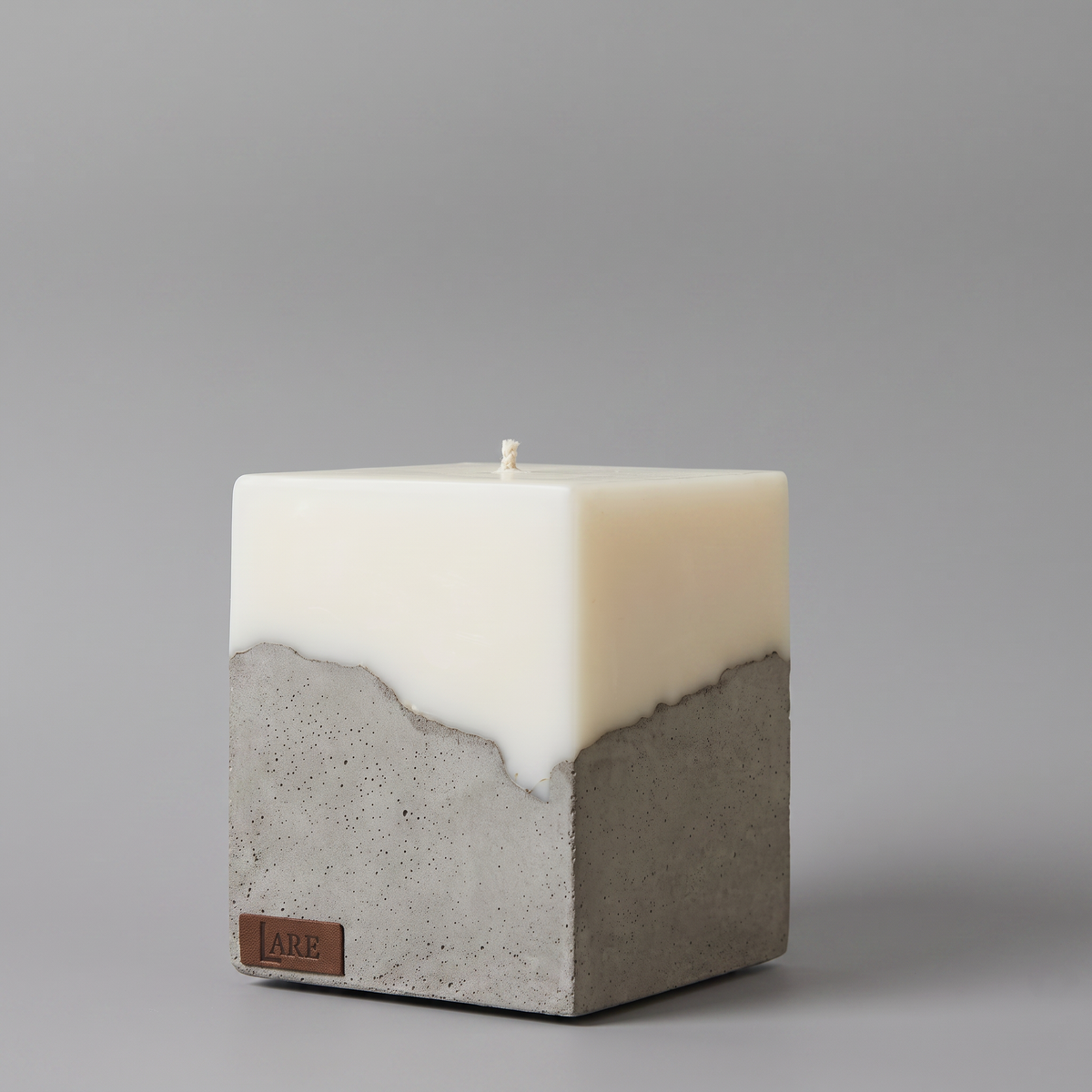 A square pillar candle with a dual-texture design, featuring a smooth soy wax top half and a textured gray concrete bottom half, with a small brown leather brand tag LARE