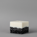 A square layered candle with a smooth matte white top half and a textured black bottom half, serving as modern bathroom decor ideas on a plain gray studio background.