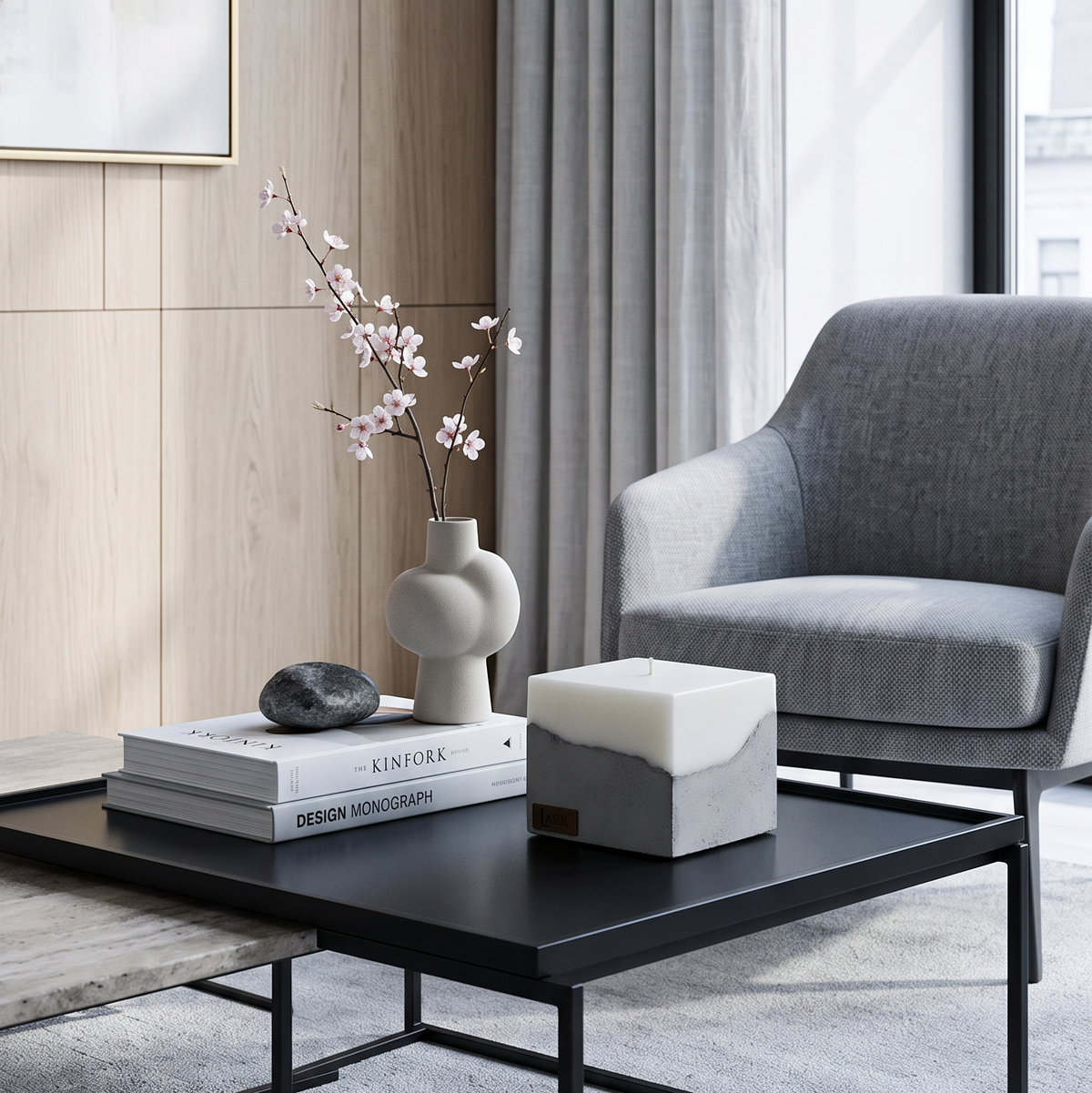 GREY sculptural soy wax candle, bringing the elegance of luxury candles to modern interiors. A modern living room scene features a black coffee table with a square candle in a white and gray marbled container, styled alongside a gray armchair, a ceramic vase with pink blossoms, and books.