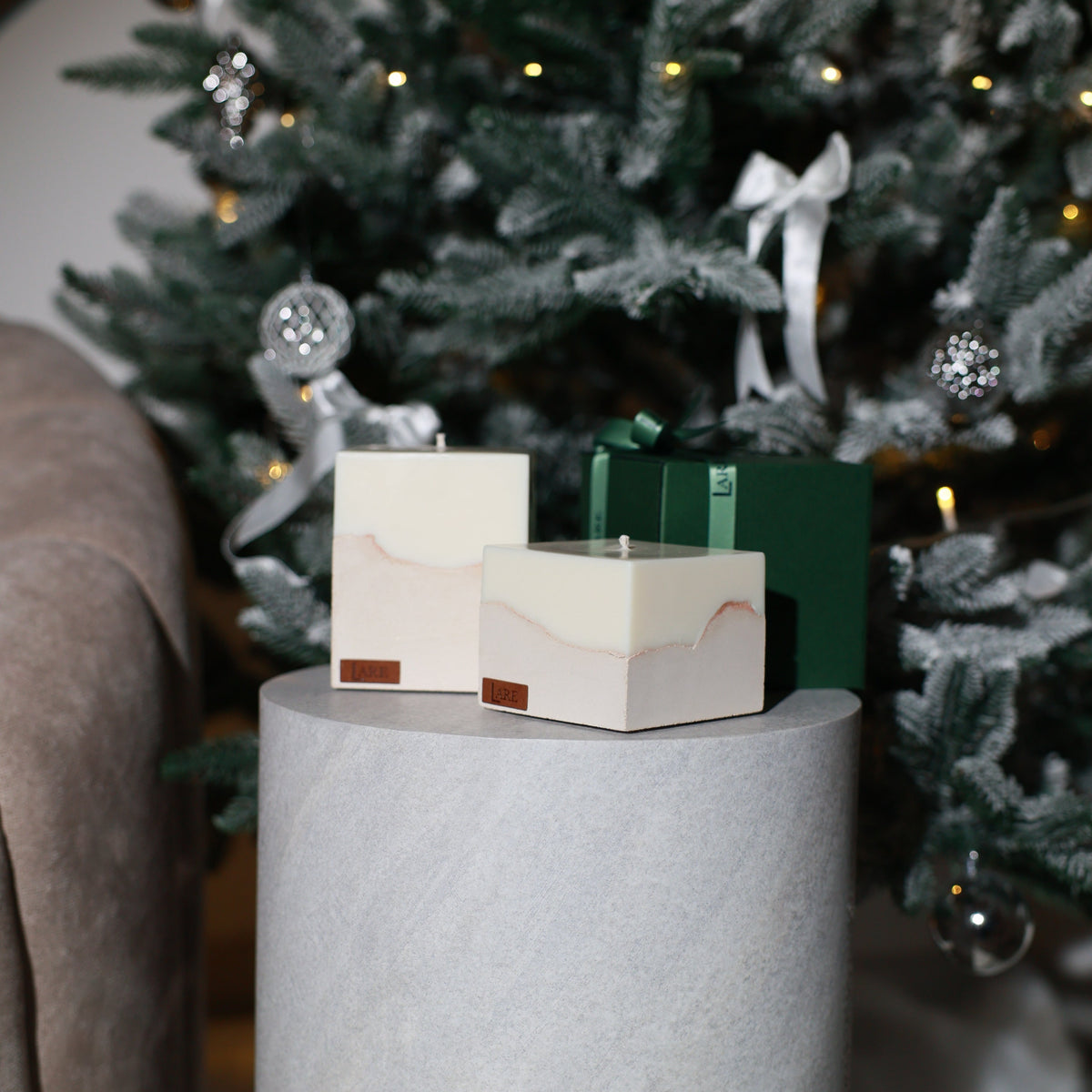 Aesthetic christmas candles, luxury-gift-ideas. Two decorative square candles in dark green packaging sit on a light gray cylindrical pedestal in front of a decorated Christmas tree.