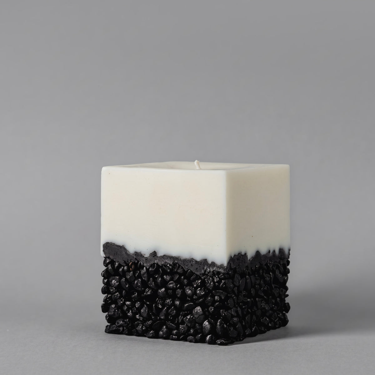 A square soy candle with a two-tone design, featuring a smooth milky top half and a textured black bottom made of small pebbles in resin, set against a solid gray background.